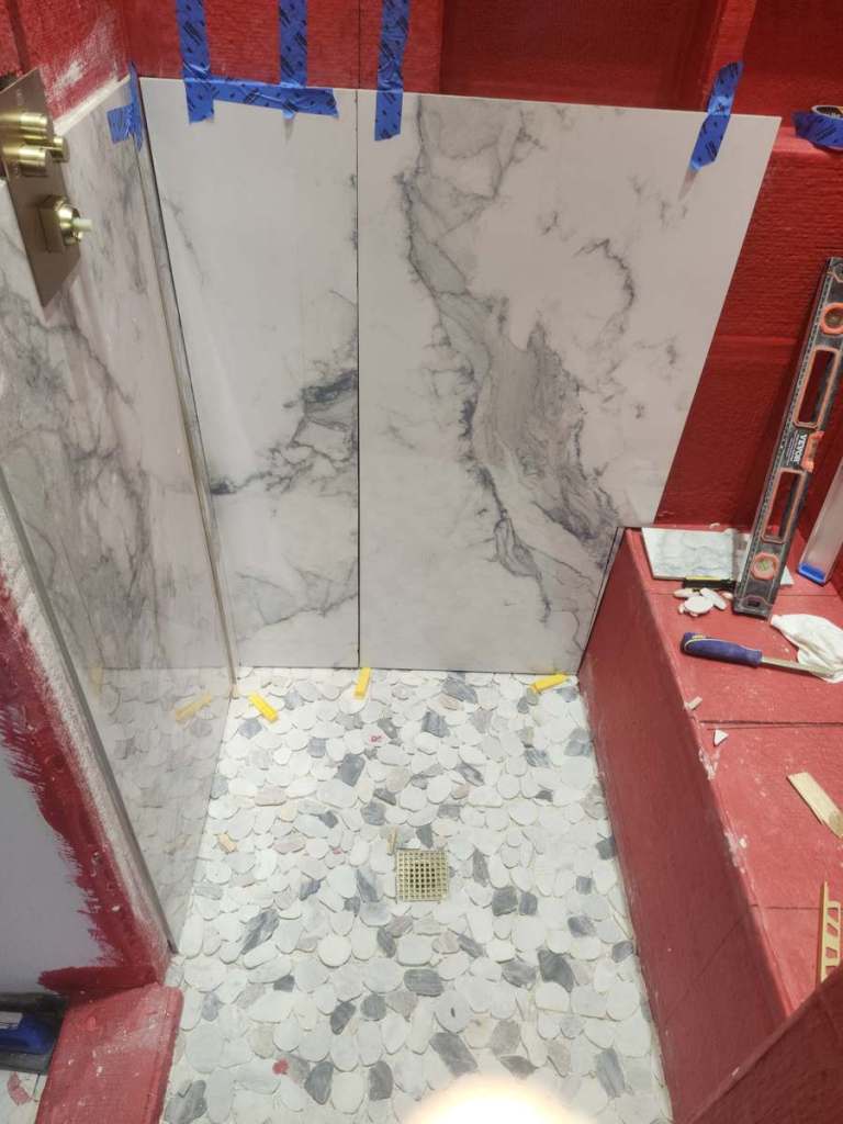 Walk-in shower in a Longmont, CO bathroom remodel: pebble stone mosaic floor, brass square drain and large-format marble-look porcelain wall tile going up over waterproofing